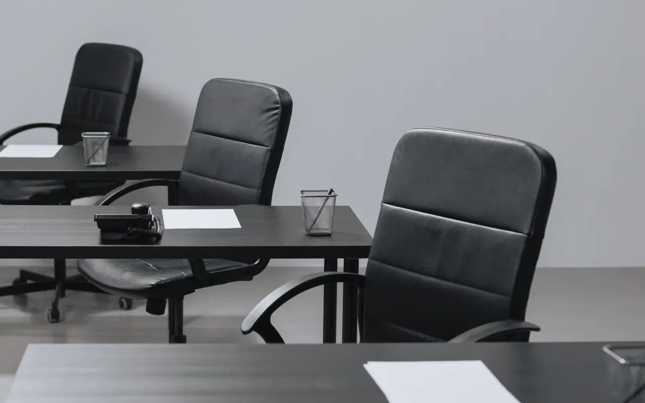 Empty black desks and office chairs in a row in front of a grey wall