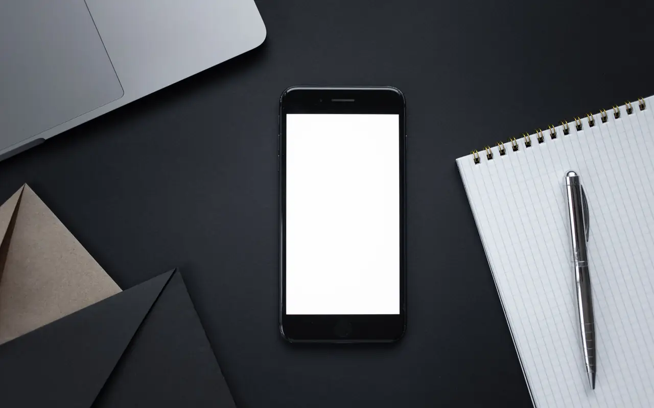 Top view of a phone with a blank screen, envelopes and a notebook with a pen on a dark desk